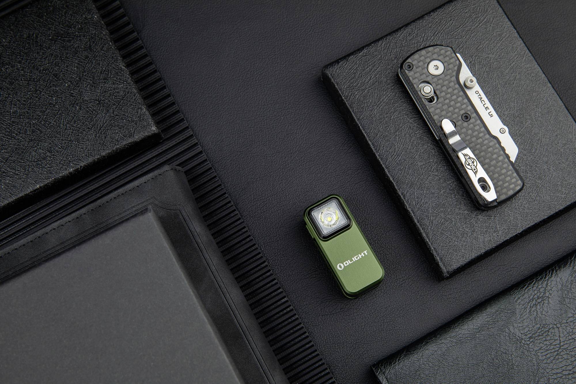 Mała latarka i składany nóż leżą obok siebie na czarnej skórzanej powierzchni. Oba przedmioty są kompaktowe i funkcjonalnie zaprojektowane.