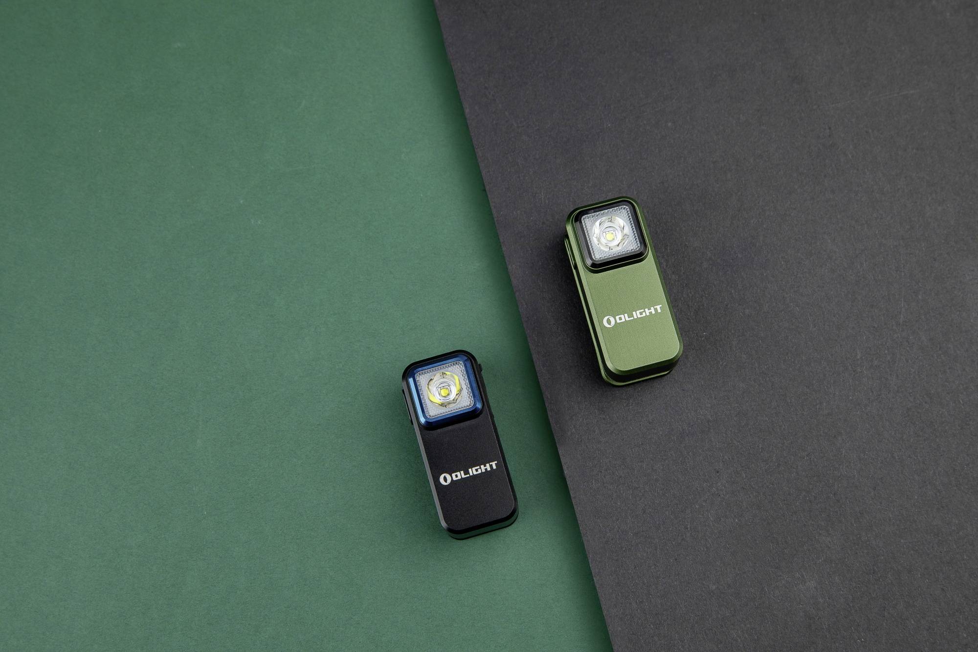 Dwie małe latarki na zielonym i czarnym tle, jedna niebieska, druga zielona, obie z logo 'Olight'.