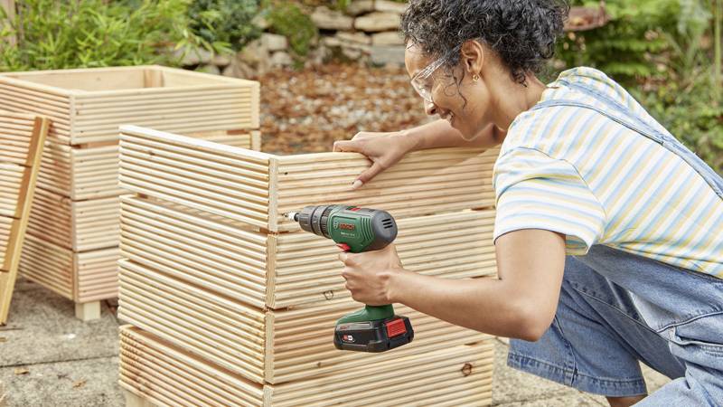 Osoba wiertli akumulatorowym wiertarką w składaną drewnianą ramę na wolnym powietrzu. W tle widoczne są kolejne elementy drewniane.
