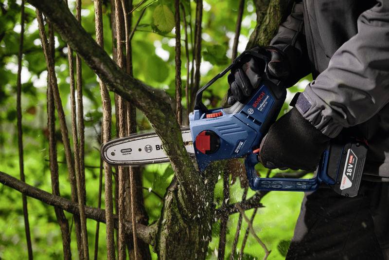 Człowiek piłuje gałęzie w lesie niebieską piłą mechaniczną. Wióry fruwają w powietrzu. Osoba ma na sobie ciemną kurtkę i rękawice.