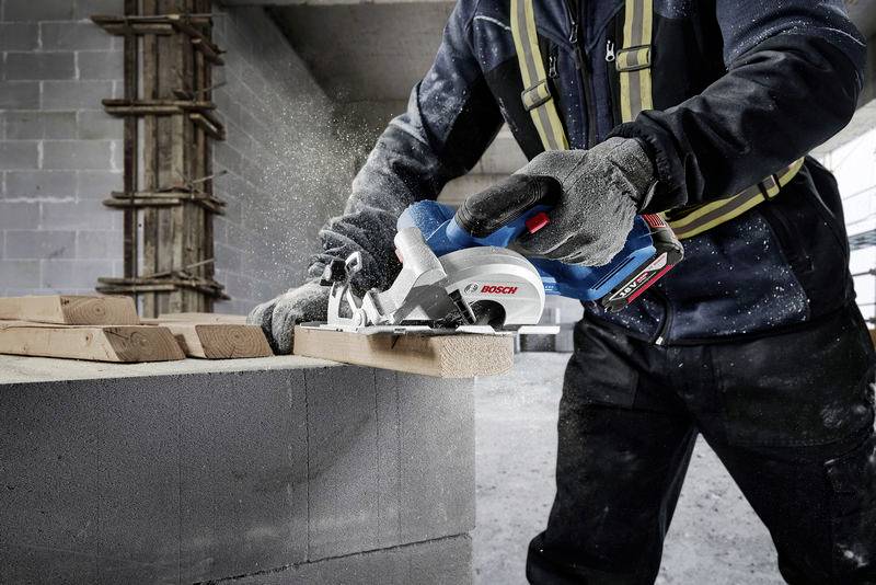 Robotnik budowlany przecina deskę drewnianą piłą ręczną Bosch na betonowej ścianie. Podczas cięcia dookoła unosi się pył.