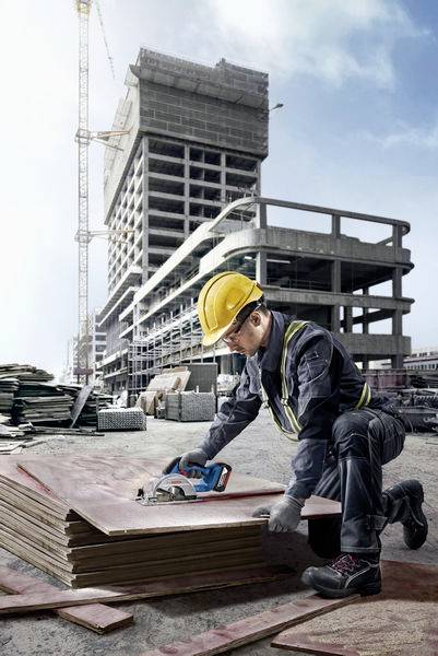 Robotnik budowlany w kasku i ubraniu ochronnym używa elektronarzędzia do cięcia desek na placu budowy, gdzie w tle widać częściowo wzniesiony budynek.