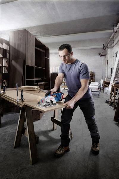 Rzemieślnik w warsztacie przecina drewno elektryczną piłą tarczową na stole warsztatowym. W tle widoczne są regały i narzędzia.