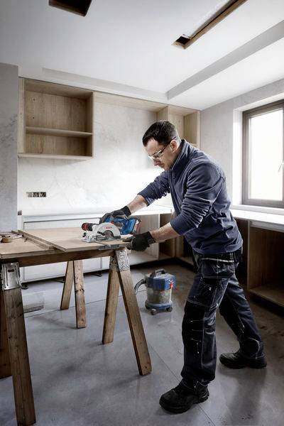 Mężczyzna pracuje w warsztacie przy desce drewnianej, używając elektrycznej piły tarczowej. W tle widoczne są okna i półki.