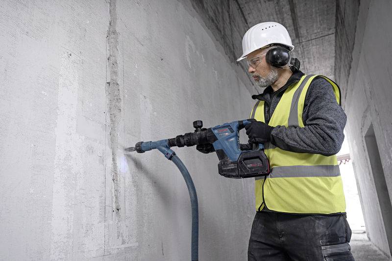 Robotnik budowlany w kasku i ubraniu ochronnym używa młotowiertarki elektrycznej, aby wiercić w betonowej ścianie.