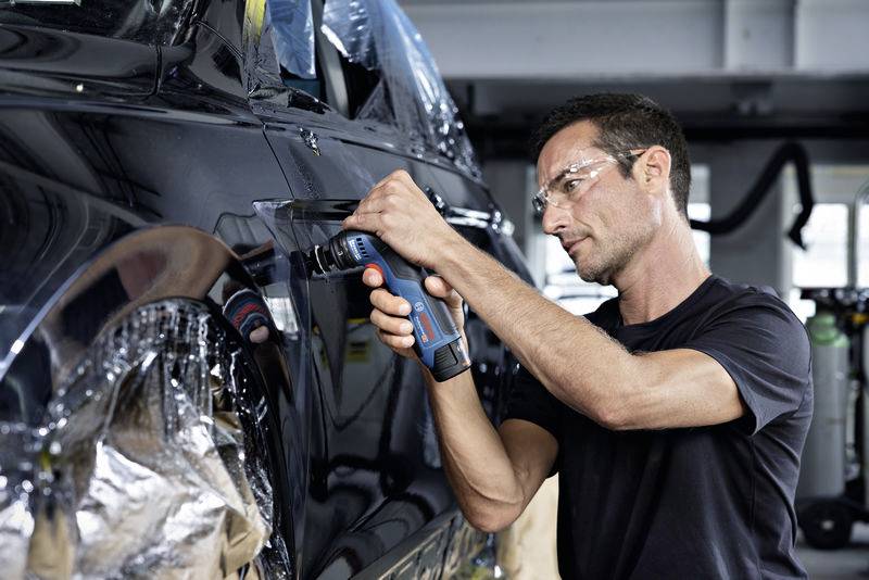 Mężczyzna w okularach ochronnych pracuje elektronarzędziem przy czarnym samochodzie w warunkach warsztatowych.