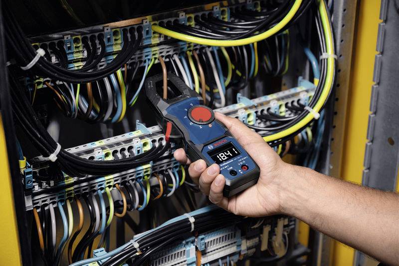 Dłoń trzyma cyfrowy przyrząd pomiarowy i mierzy natężenie prądu w rozdzielnicy elektrycznej z wieloma kablami i połączeniami.