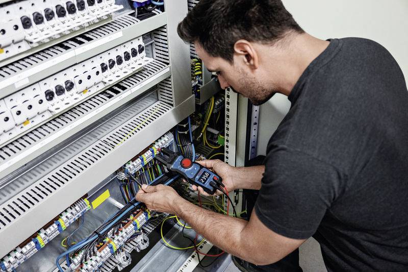 Mężczyzna naprawia elektryczną szafę rozdzielczą. Trzyma przyrząd pomiarowy i sprawdza połączenia kablowe.