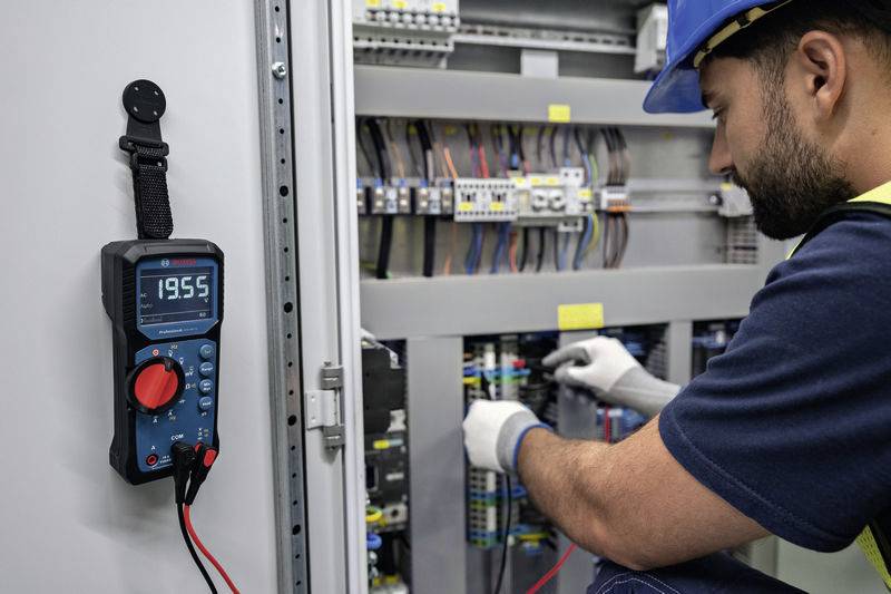 Elektryk w kasku ochronnym pracuje przy rozdzielnicy elektrycznej. Multimetr wyświetla '19,55'. Sprawdza i podłącza instalacje.