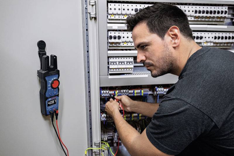 Mężczyzna pracuje skupiony przy otwartej szafce sterowniczej; multimetr wyświetla '408'. Fokus na precyzyjną konserwację elektryczną.