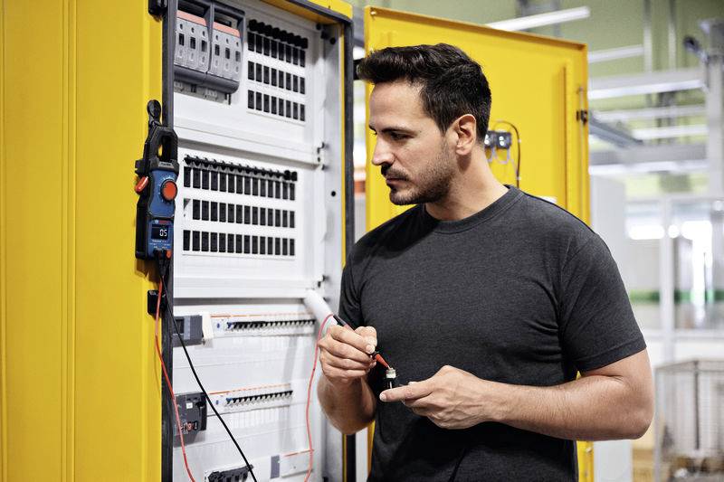 Mężczyzna sprawdza otwarty żółty rozdzielnik elektryczny za pomocą przyrządu pomiarowego. Ma na sobie ciemną koszulkę i jest całkowicie skupiony na wykonywanej pracy.