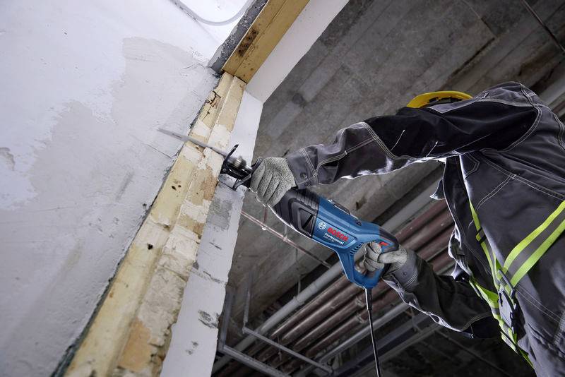 Osoba ubrana w odzież ochronną używa elektrycznej piły do cięcia płyty gipsowo-kartonowej pod sufitem.