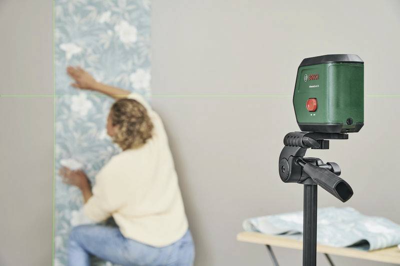 Osoba tapetuje ścianę wzdłuż zielonej linii laserowej, która jest rzutowana przez narzędzie laserowe zamontowane na statywie.