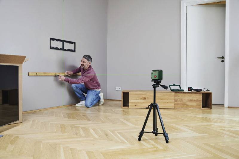 Osoba montuje uchwyt ścienny za pomocą poziomicy i lasera w pomieszczeniu z drewnianą podłogą, w tle widać statyw z kamerą.