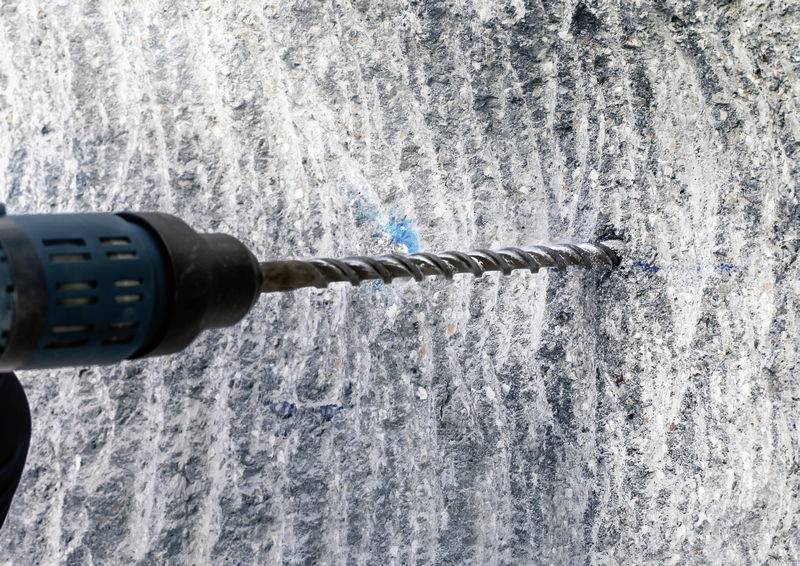 Wiertło obrabia szorstką powierzchnię kamienną, ślady procesu wiercenia są widoczne. Nacisk położony jest na końcówkę wiertła.