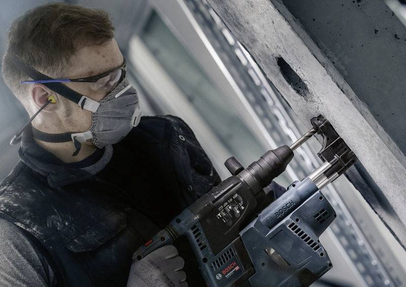 Mężczyzna w okularach ochronnych i masce wierce elektryczną wiertarką w betonową ścianę. Widoczne są środki bezpieczeństwa.