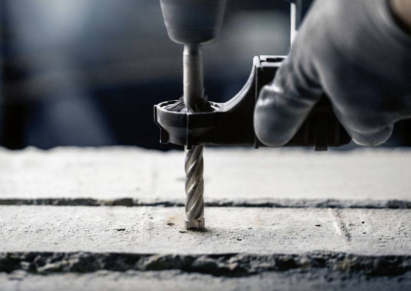 Osoba wiertarką elektryczną wykonuje otwór w betonowej powierzchni. Widoczne są rękawice robocze oraz wiertło spiralne.