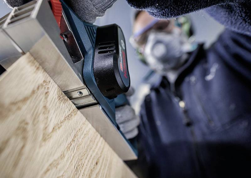 Osoba pracuje niebieską piłą elektryczną na drewnianym belkowaniu, nosi okulary ochronne i maskę przeciwpyłową.