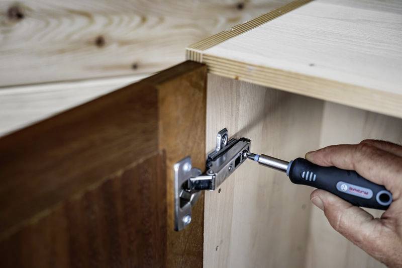 Ręka dokręca wkrętakiem śruby zawiasu drzwi drewnianej szafki.
