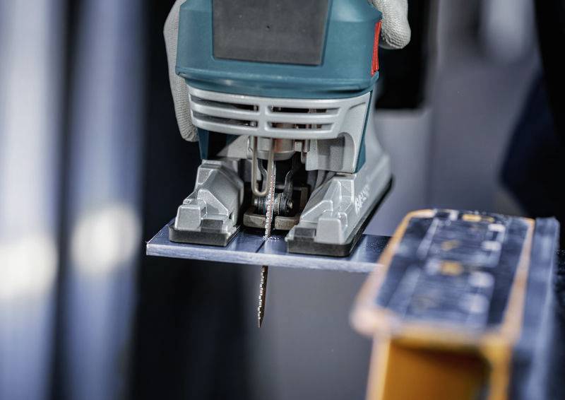 Elektryczna wyrzynarka przecina kawałek metalu. Rękawica trzyma piłę mocno.