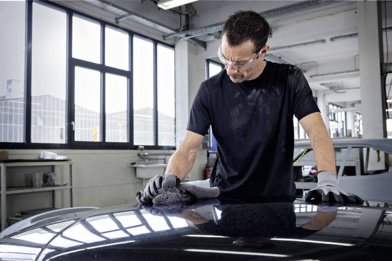 Mężczyzna w warsztacie poleruje samochód szmatką, nosi okulary ochronne i rękawice.