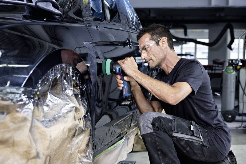 Mężczyzna poleruje czarny samochód maszyną polerską w warsztacie. Nosi okulary ochronne i czarną odzież roboczą.