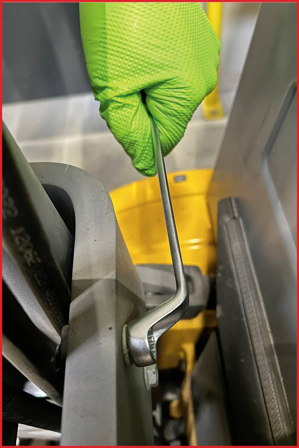 Dłoń w zielonym rękawicu obsługuje metalową dźwignię przy maszynie, dokonując regulacji lub uruchamiając jakiś mechanizm.