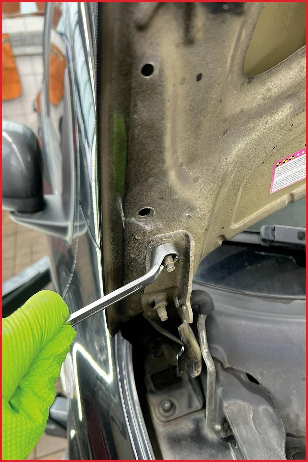 Zielona dłoń w rękawicy przykręca wkręt kluczem imbusowym w zawiasie drzwi samochodu.