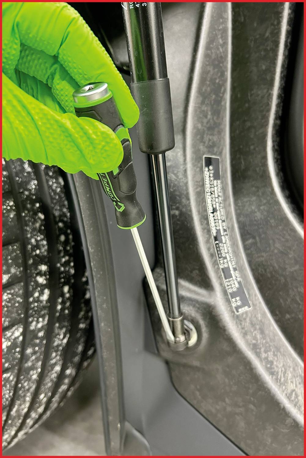 Dłoń w zielonym rękawicy trzymająca wkrętak, który mocuje śrubę w metalowym elemencie pojazdu.