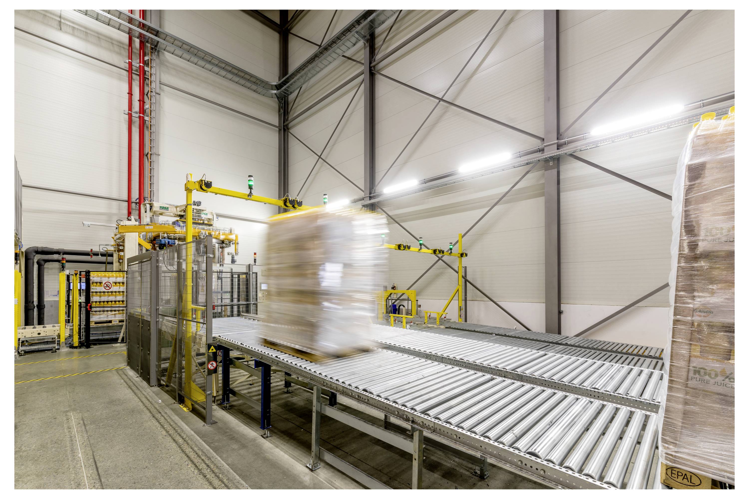 Zdjęcie magazynu przedstawia zapakowaną paletę na systemie przenośnikowym, wskazujące na zautomatyzowany transport towarów w środowisku przemysłowym.