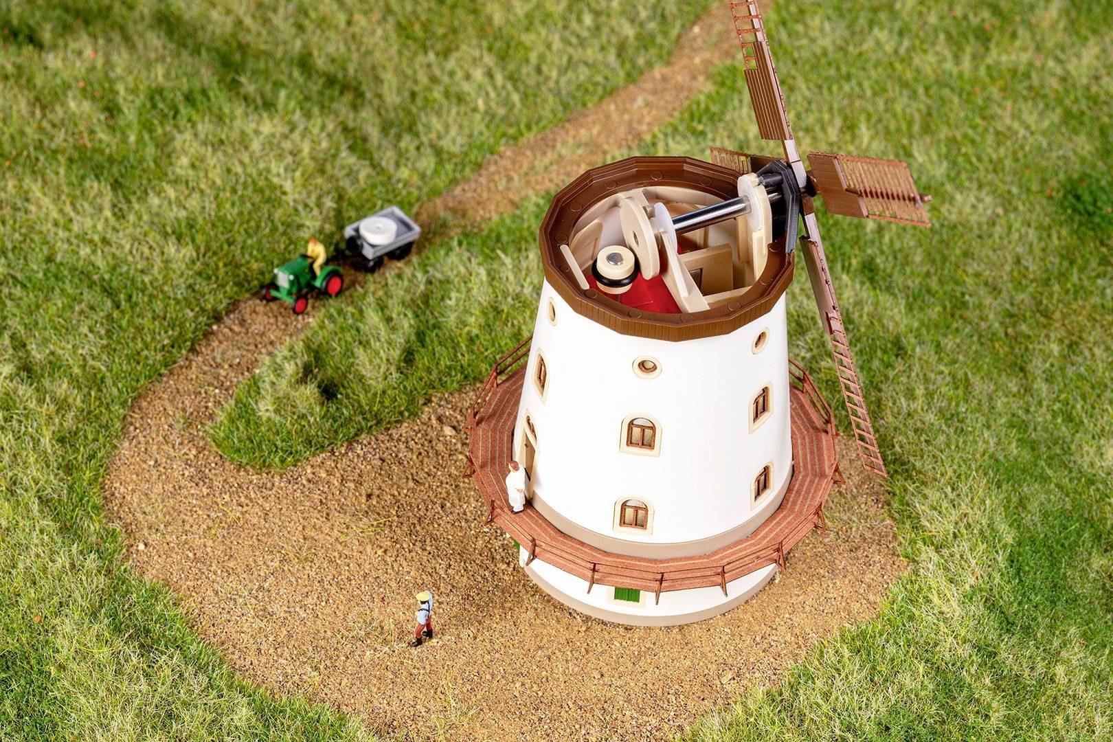 Zabawkowy wiatrak stoi na trawiastym podłożu. Mały traktor ciągnie przyczepę po drodze obok niego.