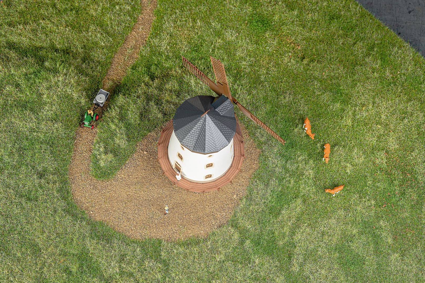 Zdjęcie lotnicze miniaturowej wiatraczki na zielonej trawie, z widoczną ścieżką, małym traktorem i trzema pomarańczowymi figurkami przedstawiającymi zwierzęta.