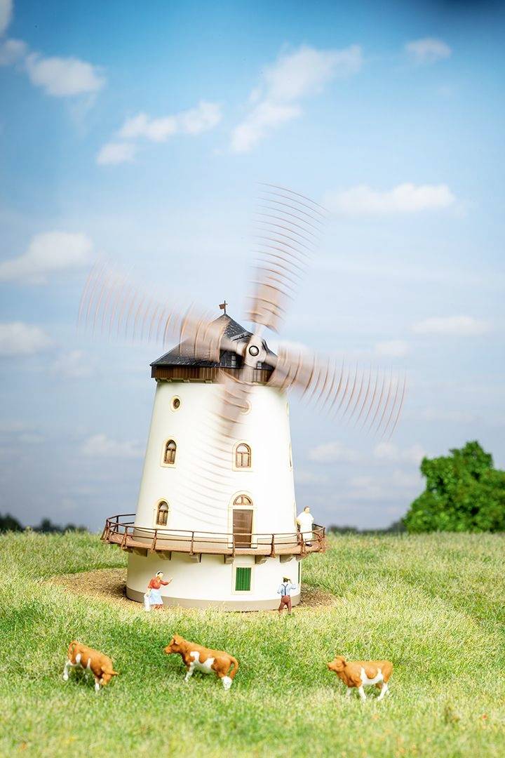 Miniaturowy model wiatraka stoi na łące, otoczony przez cztery małe krowy. Niebo jest błękitne, z nielicznymi chmurami.