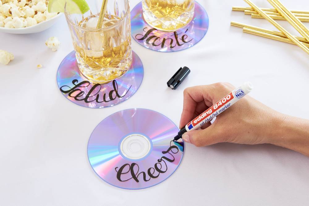 Ręka opisuje błyszczące płyty CD markerem, które służą jako podstawki pod napoje. Dwa szklanki z napojami stoją na już opisanych płytach.