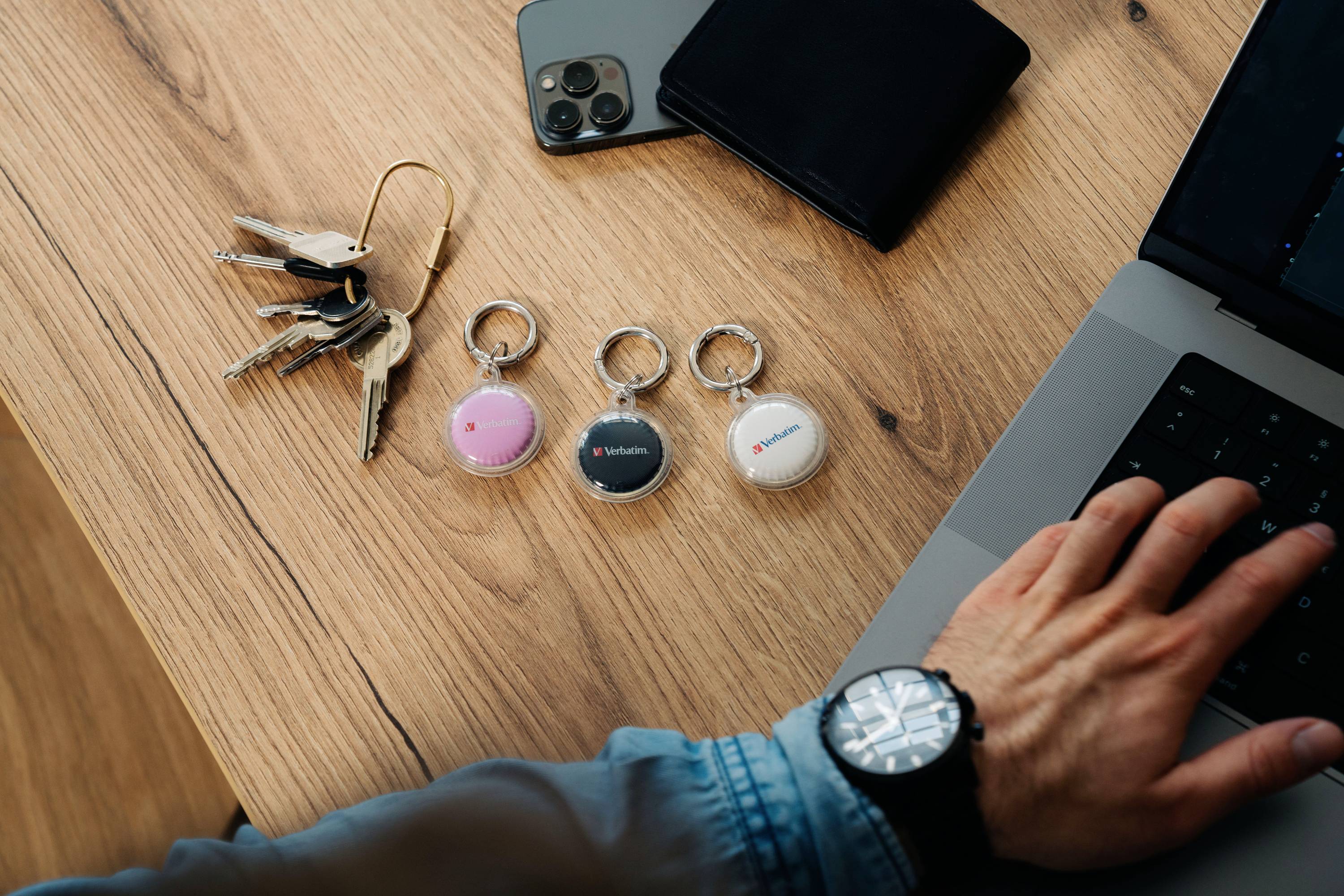 Dłoń stuka w laptopa obok smartfona, portfela i trzech zawieszek z logo marek na drewnianym stole.