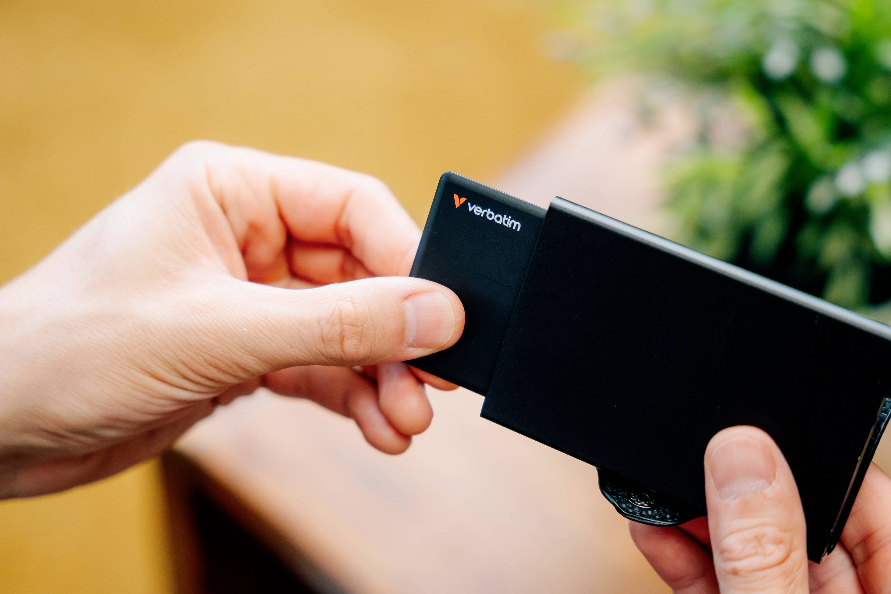 Osoba trzyma czarną kartę pamięci firmy Verbatim i wkłada ją do czarnego urządzenia w scenie we wnętrzu.