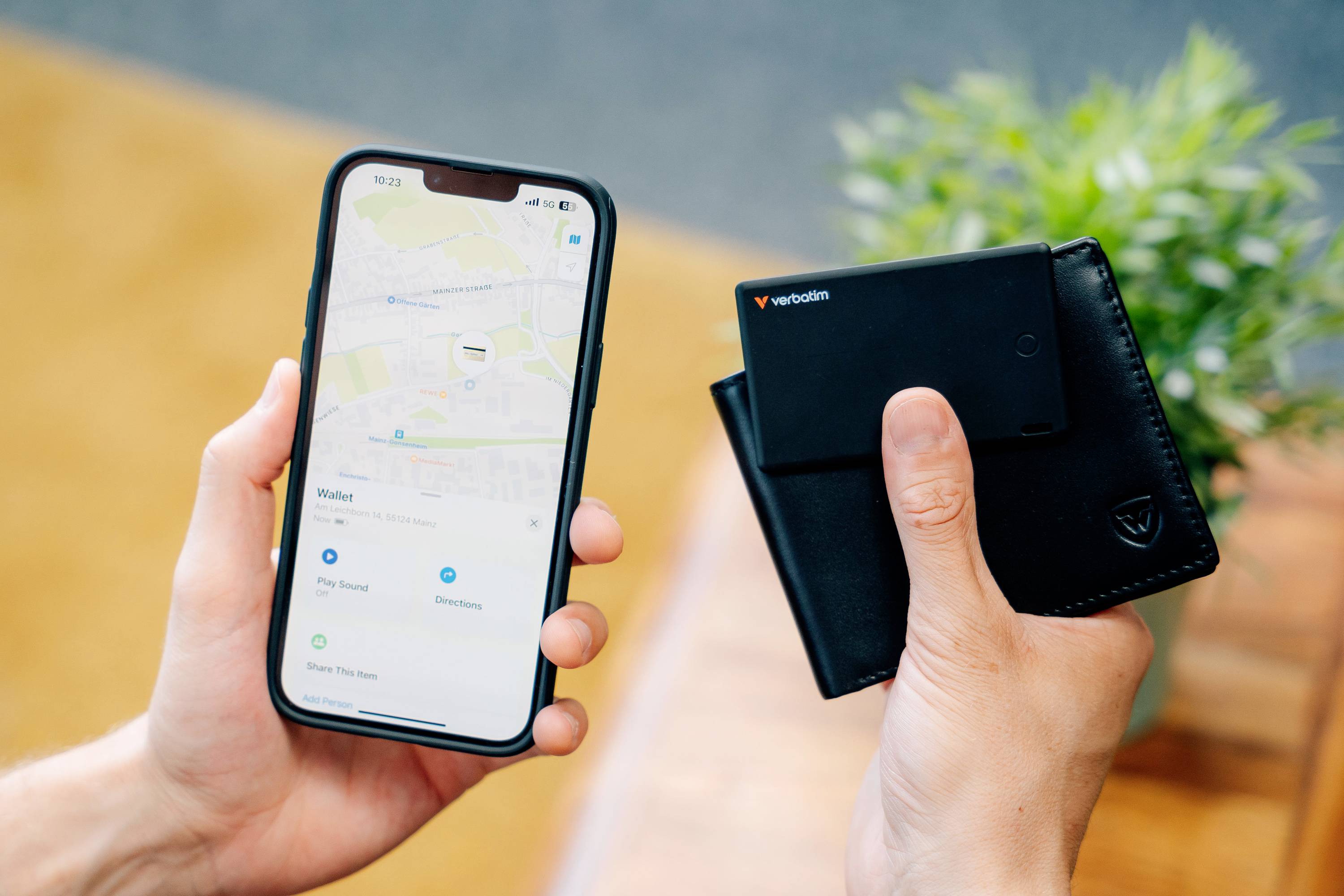 Osoba trzyma smartfon z aplikacją map w jednej ręce i czarny portfel w drugiej, w tle widać roślinę.
