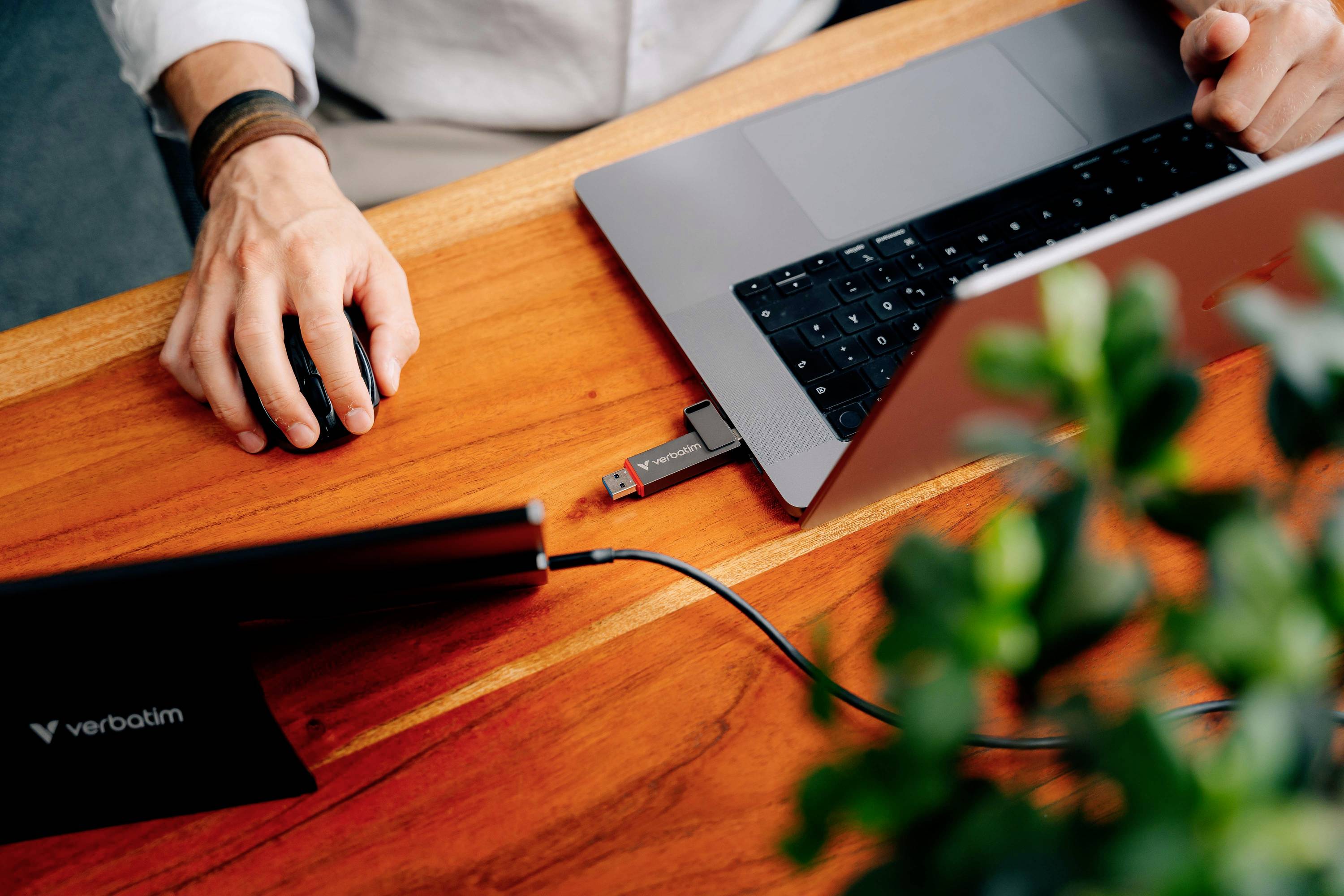 Osoba pracuje na laptopie przy drewnianym stole, podłączony pendrive, prawa ręka na myszce. Na pierwszym planie niewyraźna roślina.