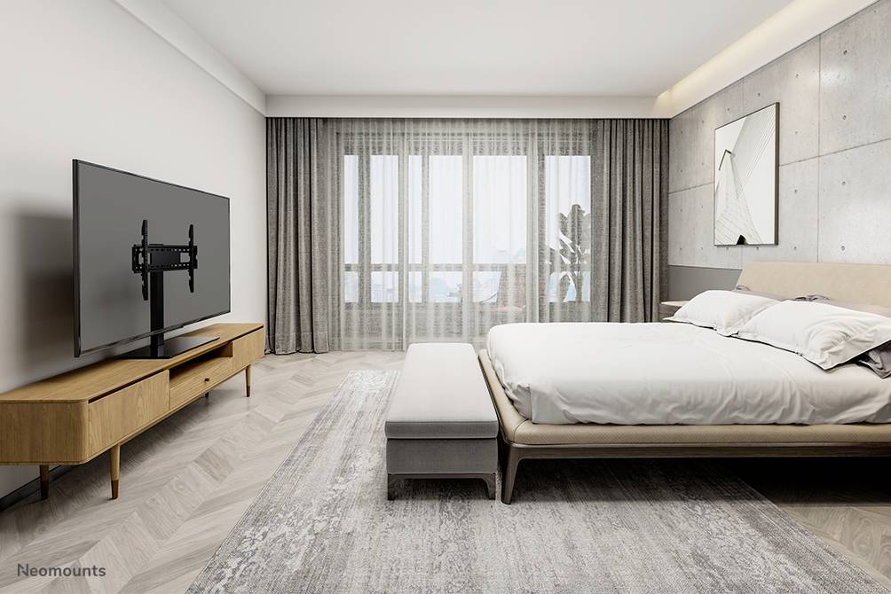 Nowoczesna sypialnia z dużym łóżkiem, drewnianym stolikiem nocnym i płaskim telewizorem na ścianie. W tle jasne okna.