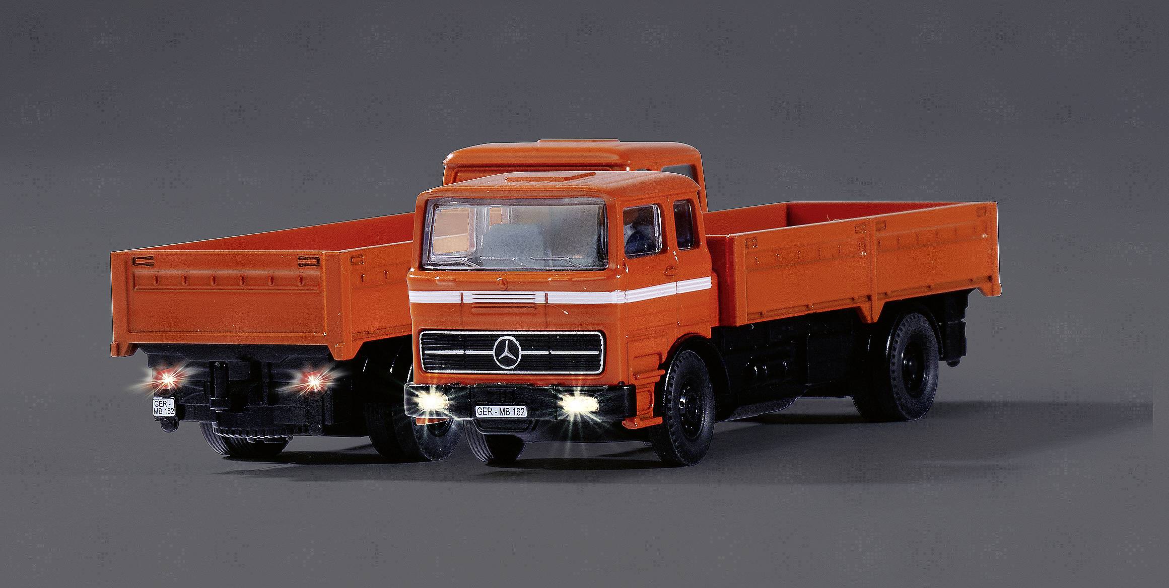 Dwa pomarańczowe ciężarówki stoją obok siebie na szarym tle. Światła są włączone.