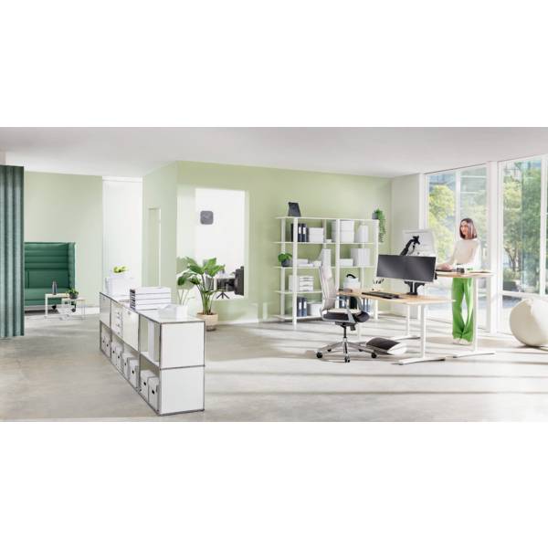 Jasne, nowoczesne biuro z zieloną ścianą akcentową, ergonomicznymi meblami, biurkiem do pracy na stojąco i dużą powierzchnią okienną. Osoba pracuje przy biurku.