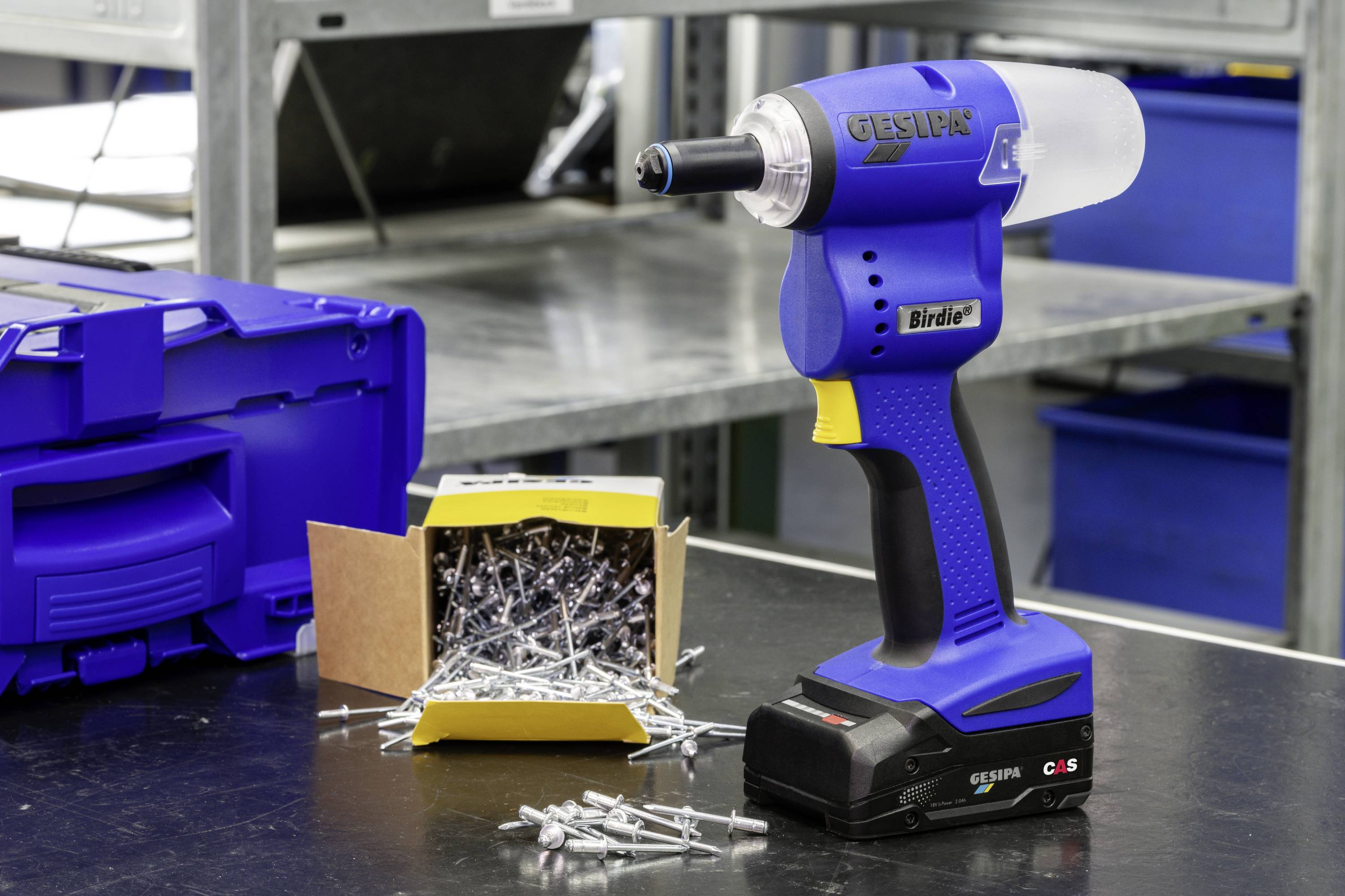 Akumulatorowa nitownica stoi pionowo na stole warsztatowym obok otwartego pudełka z nitami, a w tle widać niebieskie regały magazynowe.