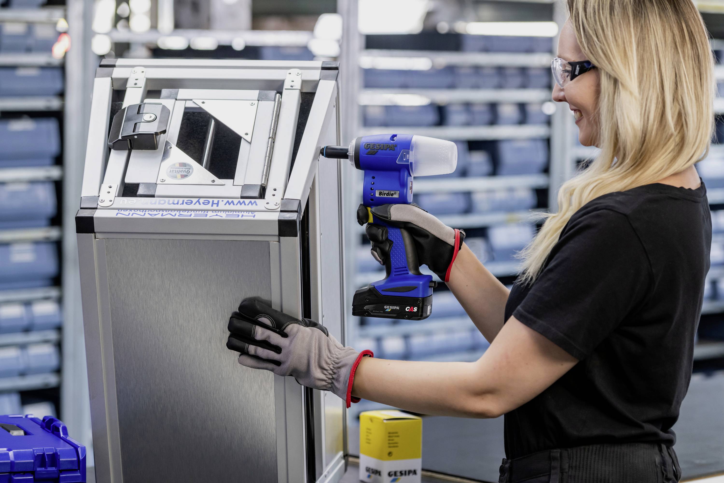 Kobieta w rękawiczkach używa elektronarzędzia na metalowej obudowie w warsztacie, wokół niej półki z niebieskimi pudełkami.