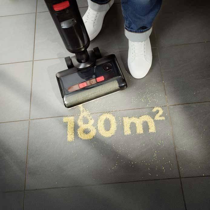 Odkurzacz usuwa okruchy z czarnej podłogi kafelkowej. Tekst na podłodze: '180 m²'. Widoczne dwie białe buty.