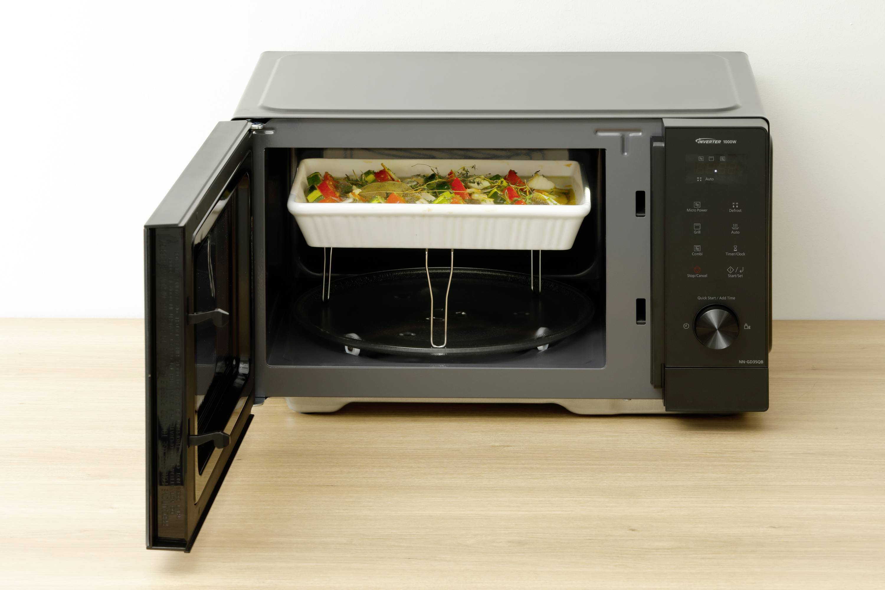 Mikrofalówka z otwartymi drzwiczkami prezentuje naczynie zapiekane z serem i warzywami w białej ceramicznej formie na obrotowym talerzu.