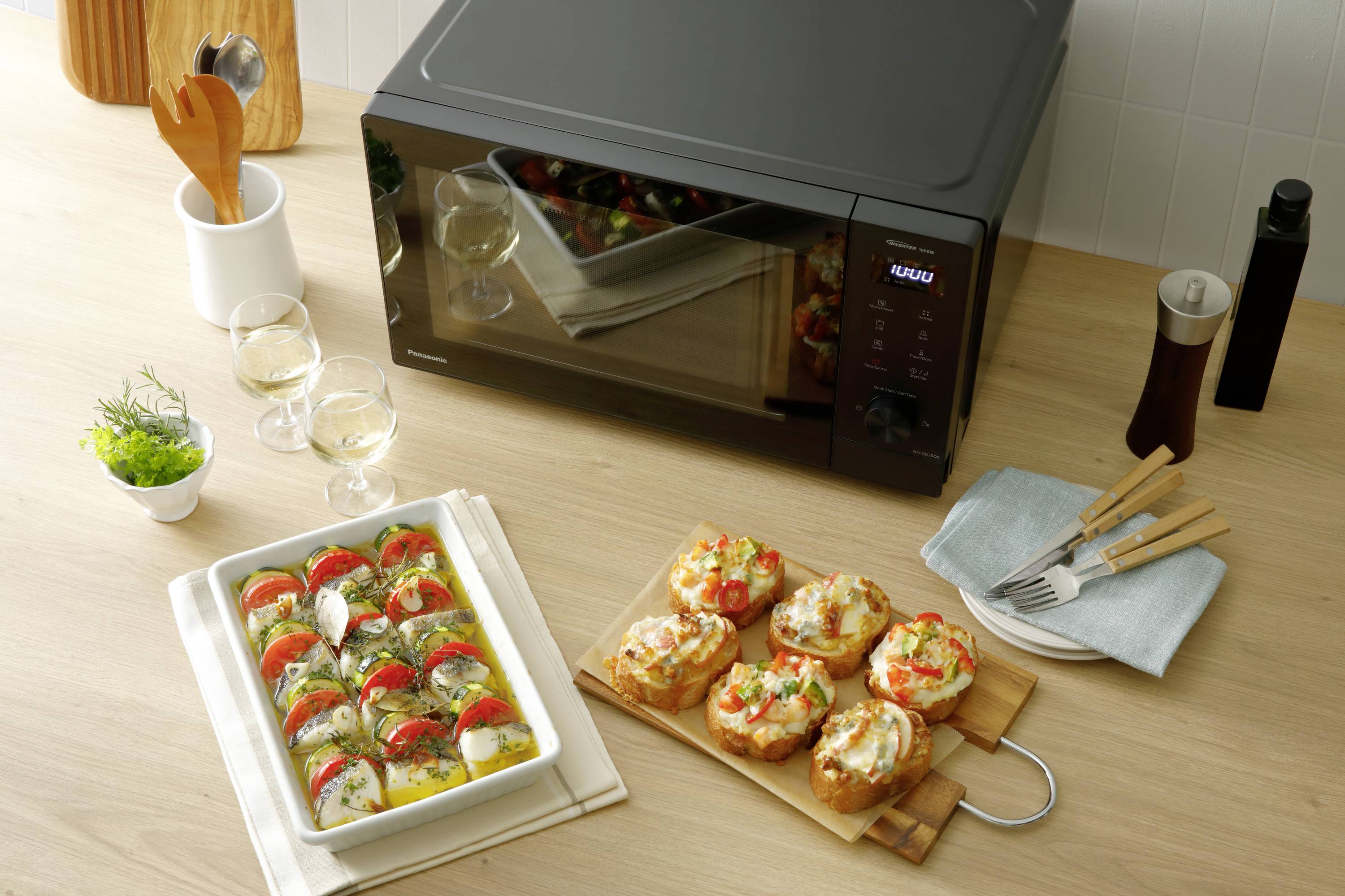 Mikrofalówka na blacie kuchennym z zapiekaną potrawą i kanapkami. Obok stoją dwa kieliszki do wina i przybory kuchenne.