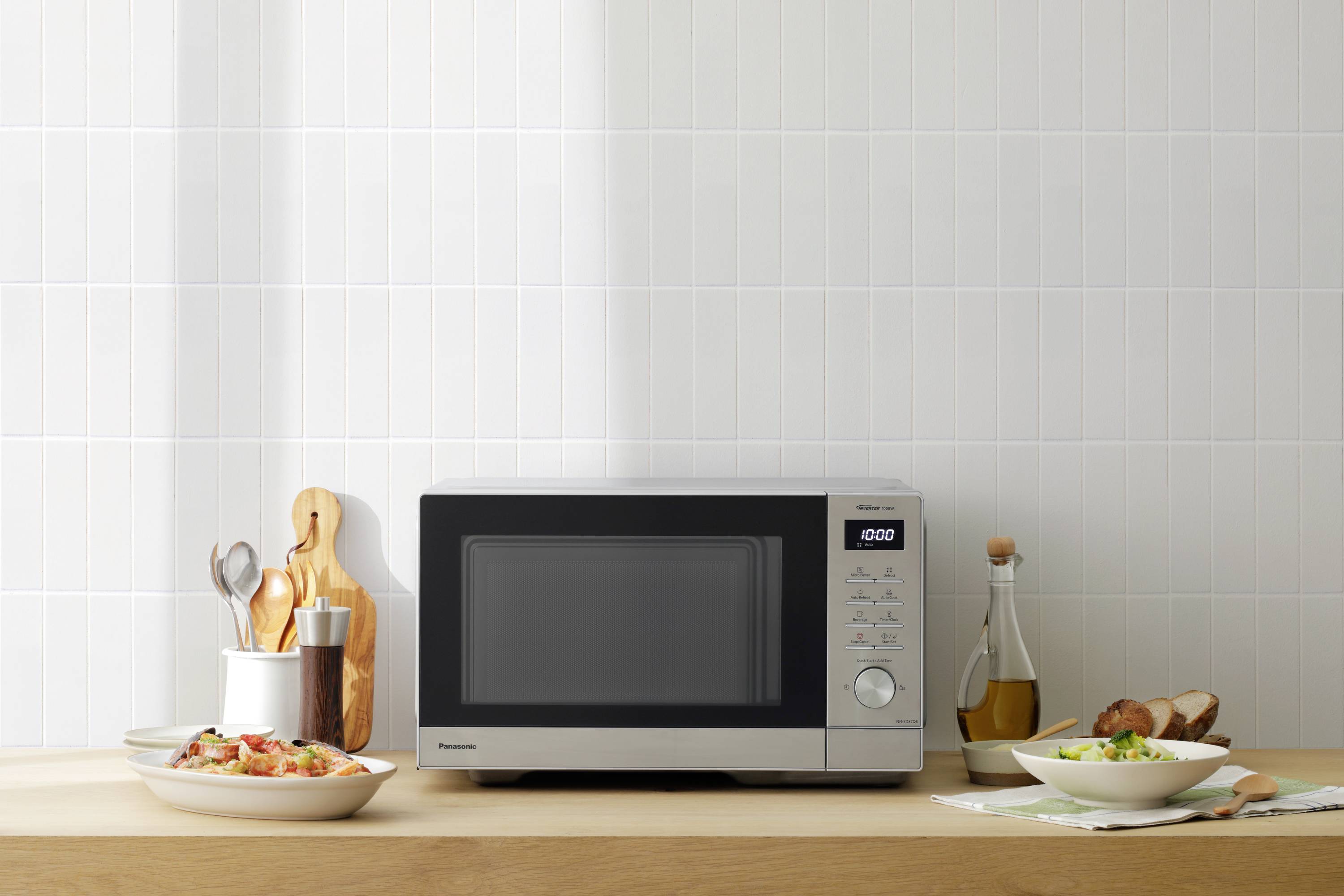 Na blacie kuchennym stoi mikrofalówka, wokół której rozłożone są dwa talerze z potrawami, bochenek chleba oraz różne przybory kuchenne.