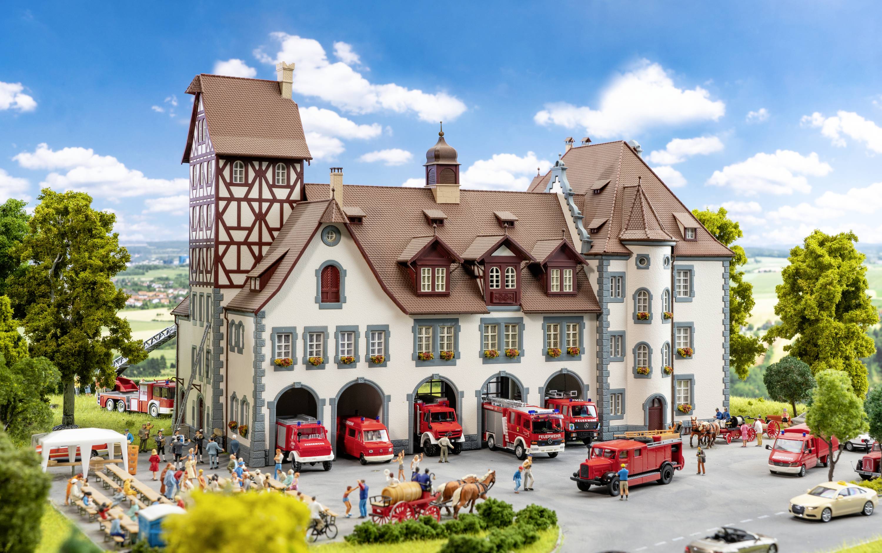 Modelarska scena kolejowa z budynkami w stylu szachulcowym, otoczonymi wozami strażackimi i miniaturowymi postaciami w wiejskim otoczeniu.