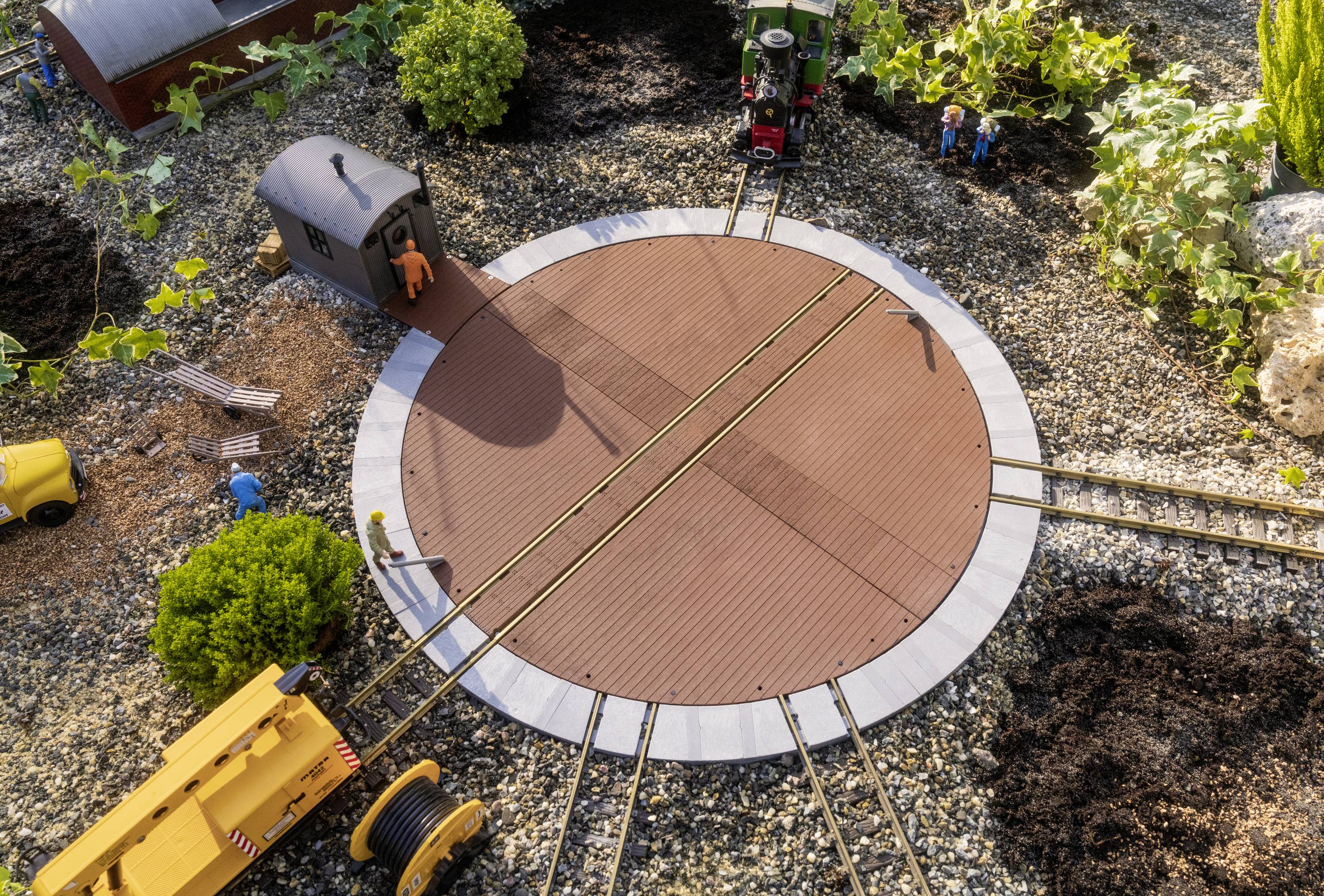 Magazyn kolejowy z obrotnicą i wieloma torami. Dwie postacie stoją obok. Rośliny i żwirowe ramy tworzą scenę.