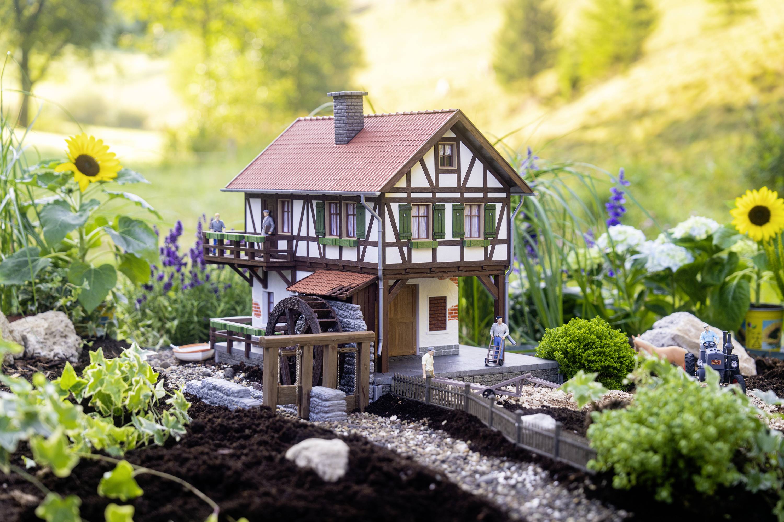 Model domu szachulcowego w miniaturowym ogrodzie z kwiatami i strumieniem, otoczony zieloną przestrzenią i słonecznikami w tle.